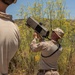A military service member in desert camouflage (MARPAT pattern) is holding and aiming a shoulder-fired weapon system, likely a man-portable air defense system (MANPADS) or anti-drone launcher, skyward. A second individual in civilian clothing is observing from the foreground. The setting appears to be a dry, scrubland environment with yellow wildflowers and hills in the background.