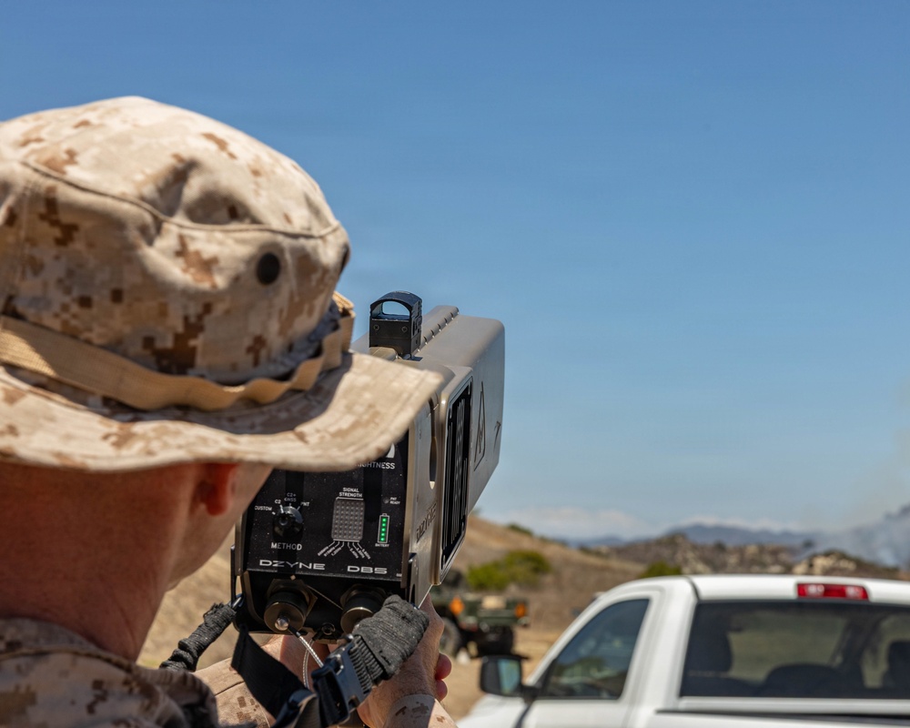 A military personnel wearing USMC MARPAT desert camouflage uniform is operating a handheld directed-energy or electronic warfare device labeled 'DZYNE DB5' in an outdoor arid environment. The device features a red dot sight on top, signal strength indicators, and multiple control settings. The setting appears to be a training exercise or field demonstration at a desert military installation.