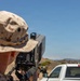 A military personnel wearing USMC MARPAT desert camouflage uniform is operating a handheld directed-energy or electronic warfare device labeled 'DZYNE DB5' in an outdoor arid environment. The device features a red dot sight on top, signal strength indicators, and multiple control settings. The setting appears to be a training exercise or field demonstration at a desert military installation.