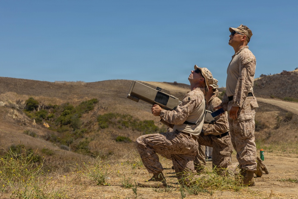Two military personnel in desert camouflage uniforms are operating what appears to be a man-portable air defense system (MANPADS) or directed-energy weapon in an outdoor training environment. One soldier is kneeling and aiming the device skyward while the other stands behind observing upward. The setting appears to be a dry, hilly terrain consistent with a military training range in a semi-arid region.