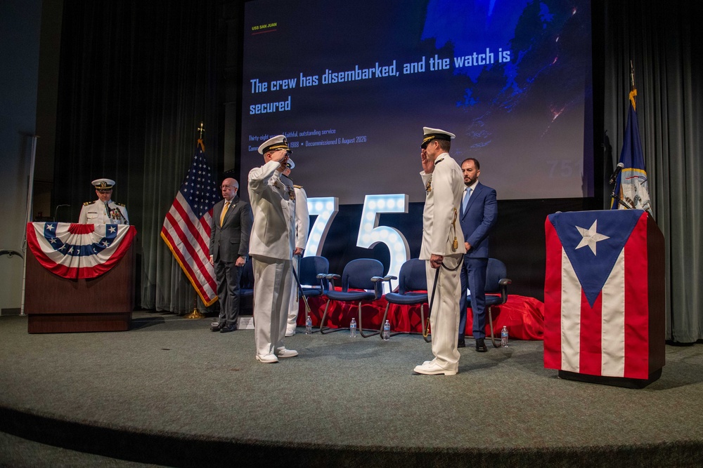 This image depicts a formal U.S. Navy decommissioning ceremony, as indicated by the projected text reading 'The crew has disembarked, and the watch is secured.' The slide references 'USS San Juan' and notes it was decommissioned on 6 August 2026 after thirty-eight years of service. Two naval officers in white dress uniforms are exchanging salutes on stage, flanked by dignitaries and flags including the U.S. flag and the Puerto Rico flag.