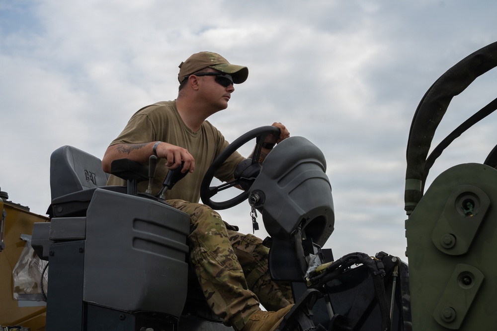 A military service member in OCP (Operational Camouflage Pattern) uniform is operating what appears to be a Caterpillar (CAT) construction or utility vehicle. The individual is wearing a multicam cap, sunglasses, and an olive drab t-shirt, seated at the controls of the equipment. The image is taken from a low angle against a partly cloudy sky.