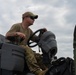 A military service member in OCP (Operational Camouflage Pattern) uniform is operating what appears to be a Caterpillar (CAT) construction or utility vehicle. The individual is wearing a multicam cap, sunglasses, and an olive drab t-shirt, seated at the controls of the equipment. The image is taken from a low angle against a partly cloudy sky.