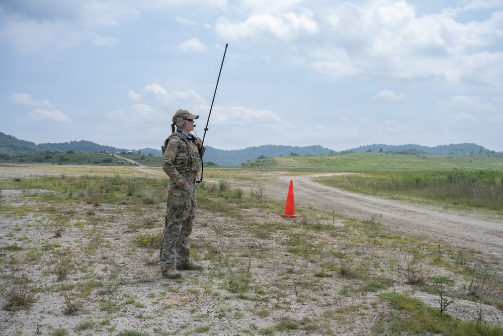 A military service member in full camouflage uniform stands on an open sandy field, holding a long-antenna radio and appearing to communicate. The setting appears to be a military training area or airfield, marked by an orange traffic cone and a dirt road in the background. Rolling green hills and a partly cloudy sky form the backdrop.