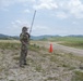 A military service member in full camouflage uniform stands on an open sandy field, holding a long-antenna radio and appearing to communicate. The setting appears to be a military training area or airfield, marked by an orange traffic cone and a dirt road in the background. Rolling green hills and a partly cloudy sky form the backdrop.