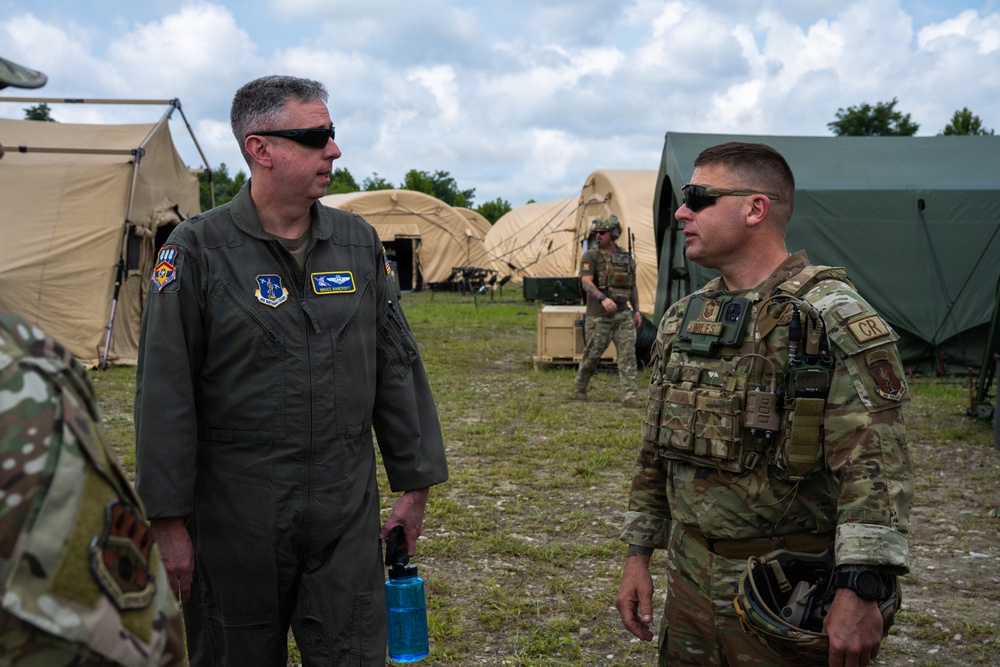 Two U.S. Air Force personnel are engaged in conversation at what appears to be a forward operating base or field training exercise. The individual on the left is wearing an olive green flight suit with unit patches and pilot wings, while the individual on the right is in full MultiCam combat gear with a plate carrier vest. Multiple tan and green military tents are visible in the background along with additional personnel.