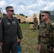 Two U.S. Air Force personnel are engaged in conversation at what appears to be a forward operating base or field training exercise. The individual on the left is wearing an olive green flight suit with unit patches and pilot wings, while the individual on the right is in full MultiCam combat gear with a plate carrier vest. Multiple tan and green military tents are visible in the background along with additional personnel.