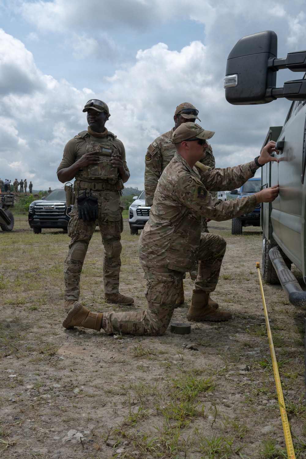Three military personnel in MultiCam/OCP camouflage uniforms are gathered near a military vehicle in an outdoor field setting. One soldier is kneeling and appears to be working on or inspecting the side of a large vehicle, while another stands nearby wearing a plate carrier vest. A third individual stands in the background, and additional personnel and vehicles are visible in the distance.
