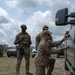 Three military personnel in MultiCam/OCP camouflage uniforms are gathered near a military vehicle in an outdoor field setting. One soldier is kneeling and appears to be working on or inspecting the side of a large vehicle, while another stands nearby wearing a plate carrier vest. A third individual stands in the background, and additional personnel and vehicles are visible in the distance.