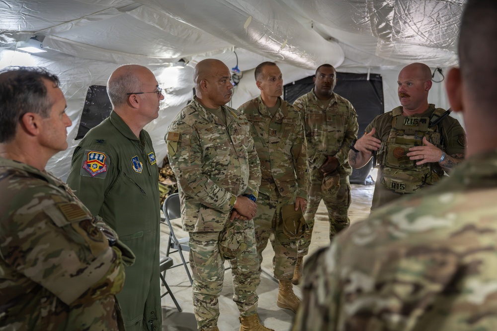 A group of military personnel are gathered inside a large white tent structure for what appears to be a briefing or discussion. One individual wearing a tactical vest with visible markings appears to be addressing the group, while others listen attentively. The group includes personnel in both OCP (Operational Camouflage Pattern) uniforms and one individual in an olive green flight suit with unit patches.