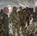 A group of military personnel are gathered inside a large white tent structure for what appears to be a briefing or discussion. One individual wearing a tactical vest with visible markings appears to be addressing the group, while others listen attentively. The group includes personnel in both OCP (Operational Camouflage Pattern) uniforms and one individual in an olive green flight suit with unit patches.