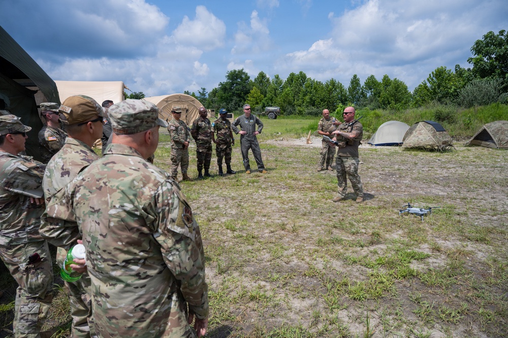 A group of military personnel in various camouflage uniforms are gathered in an outdoor field setting, appearing to receive a briefing or demonstration. A small consumer drone (likely a DJI quadcopter) is visible on the ground in the foreground, suggesting a UAS (unmanned aerial systems) training or demonstration session. The setting includes military tents, a vehicle in the background, and a treeline, consistent with a field training exercise environment.
