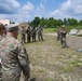 A group of military personnel in various camouflage uniforms are gathered in an outdoor field setting, appearing to receive a briefing or demonstration. A small consumer drone (likely a DJI quadcopter) is visible on the ground in the foreground, suggesting a UAS (unmanned aerial systems) training or demonstration session. The setting includes military tents, a vehicle in the background, and a treeline, consistent with a field training exercise environment.