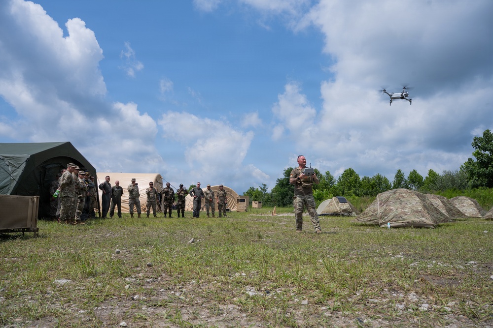 A military training exercise is taking place at an outdoor field camp where a soldier in camouflage uniform is operating a small quadcopter drone using a handheld controller. A group of approximately 12-15 military personnel in various uniforms observe from the left side near military tents. The setting appears to be a temporary field base with multiple tents, open grassy terrain, and tree lines in the background.