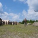 A military training exercise is taking place at an outdoor field camp where a soldier in camouflage uniform is operating a small quadcopter drone using a handheld controller. A group of approximately 12-15 military personnel in various uniforms observe from the left side near military tents. The setting appears to be a temporary field base with multiple tents, open grassy terrain, and tree lines in the background.