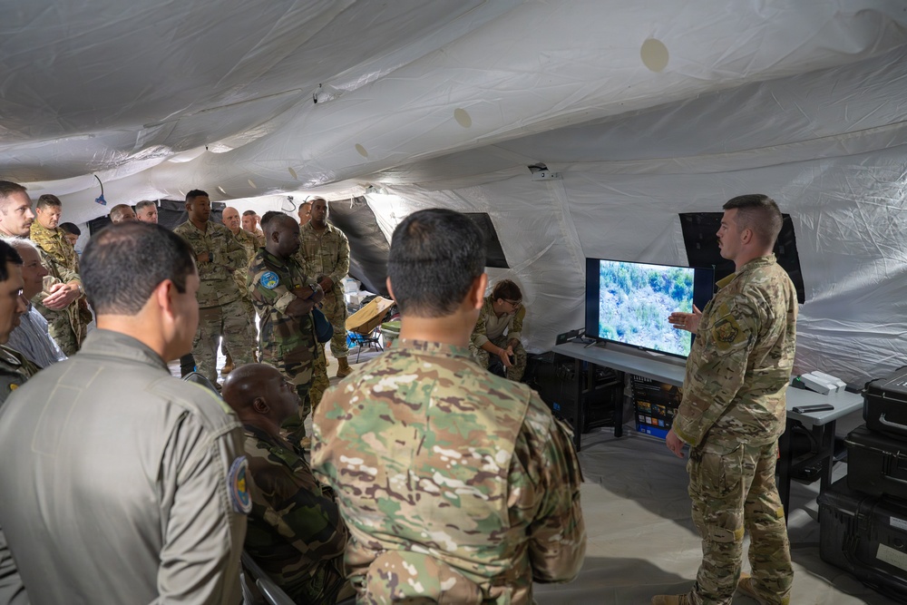 A group of military personnel in various camouflage uniforms are gathered inside a large white inflatable or canvas tent structure for what appears to be a briefing or demonstration. One soldier stands to the right presenting information displayed on a flat-screen monitor showing aerial or overhead imagery of a forested area. The audience includes personnel wearing different uniform patterns, suggesting a multinational or joint-service gathering.