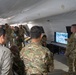 A group of military personnel in various camouflage uniforms are gathered inside a large white inflatable or canvas tent structure for what appears to be a briefing or demonstration. One soldier stands to the right presenting information displayed on a flat-screen monitor showing aerial or overhead imagery of a forested area. The audience includes personnel wearing different uniform patterns, suggesting a multinational or joint-service gathering.