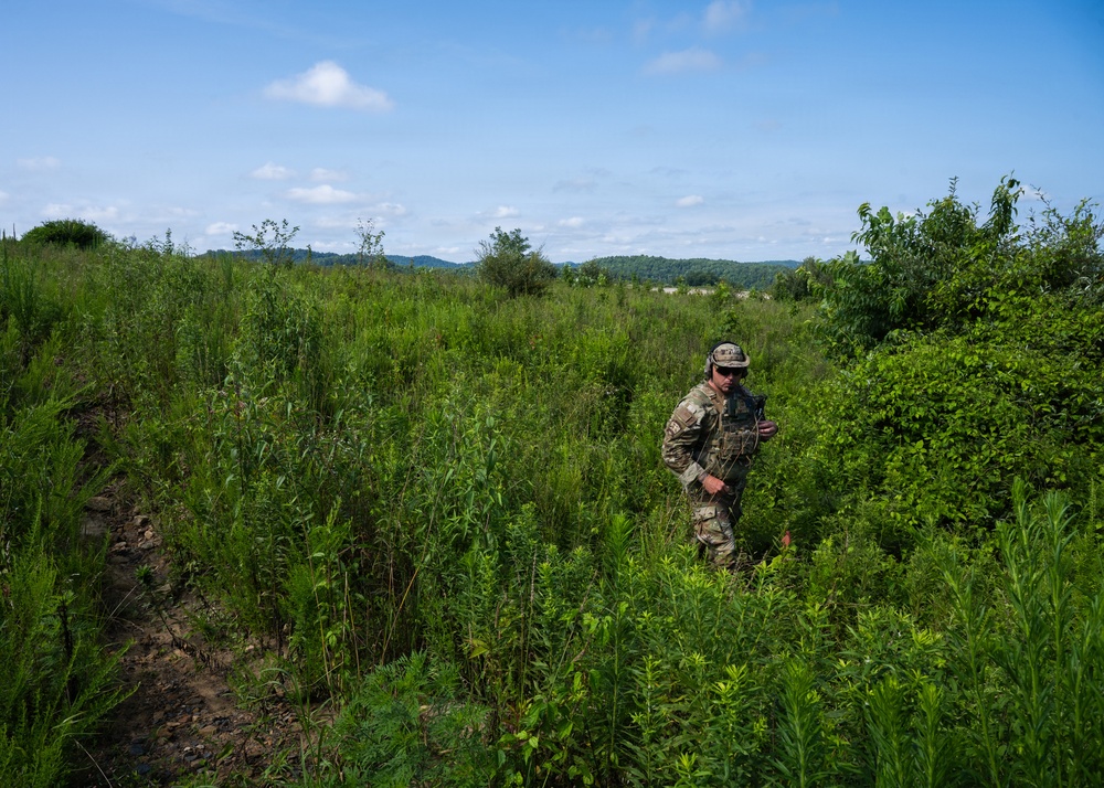 A soldier in full camouflage uniform and tactical gear navigates through dense, overgrown vegetation in what appears to be a field training exercise. The individual is moving through thick brush and tall weeds in a rural, hilly landscape under a partly cloudy sky. The setting suggests a military reconnaissance or patrol training scenario.