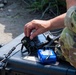 A military personnel in camouflage uniform is operating tactical radio communication equipment placed on a hard-shell protective case outdoors. The individual is handling what appears to be a field radio transceiver with multiple cables and a battery pack connected to it. The setting appears to be an outdoor field environment, suggesting a training exercise or operational deployment.