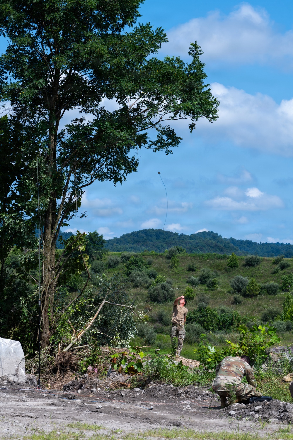 Two individuals in military camouflage uniforms are working outdoors in a cleared area near a tall tree. One person is standing and looking upward, apparently throwing or launching a weighted line into the tree, while a second person crouches nearby on the ground. The setting appears to be a rural or training area with rolling hills and vegetation in the background.