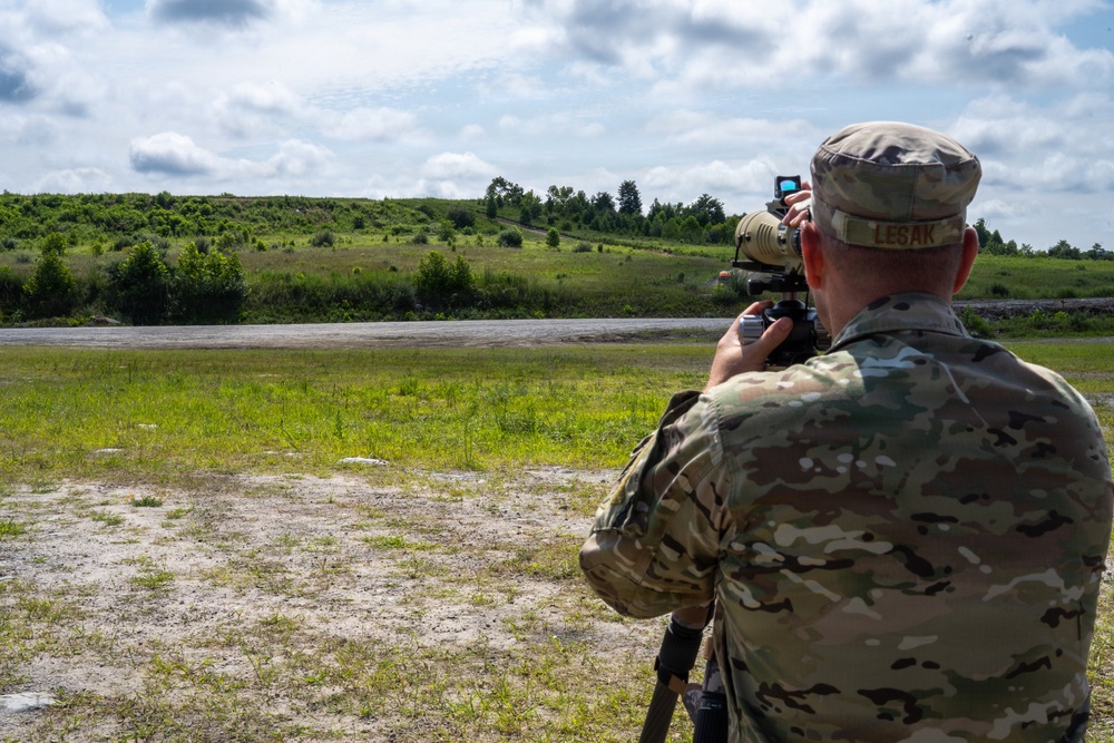 A military service member identified by the name tape 'LESAK' is using a spotting scope mounted on a tripod at what appears to be a firing range or training area. The soldier is wearing MultiCam (OCP) camouflage uniform and patrol cap, and is observing a downrange area with green hills and treelines in the background. The setting suggests a live-fire training exercise or marksmanship qualification event.