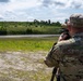 A military service member identified by the name tape 'LESAK' is using a spotting scope mounted on a tripod at what appears to be a firing range or training area. The soldier is wearing MultiCam (OCP) camouflage uniform and patrol cap, and is observing a downrange area with green hills and treelines in the background. The setting suggests a live-fire training exercise or marksmanship qualification event.