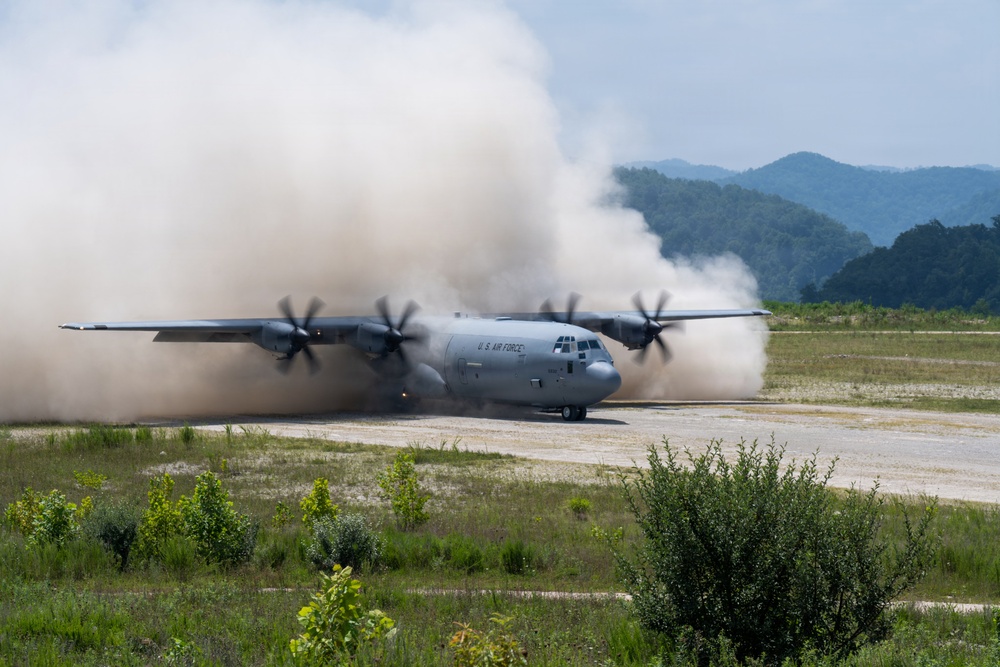 A U.S. Air Force C-130J Super Hercules military transport aircraft is operating on an unpaved or semi-prepared airstrip, generating large dust clouds from its four turboprop engines. The aircraft bears visible 'U.S. AIR FORCE' markings on its fuselage and appears to be either landing or conducting a short-field takeoff. The setting features a mountainous, heavily forested background suggesting a location in East or Southeast Asia.