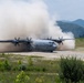 A U.S. Air Force C-130J Super Hercules military transport aircraft is operating on an unpaved or semi-prepared airstrip, generating large dust clouds from its four turboprop engines. The aircraft bears visible 'U.S. AIR FORCE' markings on its fuselage and appears to be either landing or conducting a short-field takeoff. The setting features a mountainous, heavily forested background suggesting a location in East or Southeast Asia.