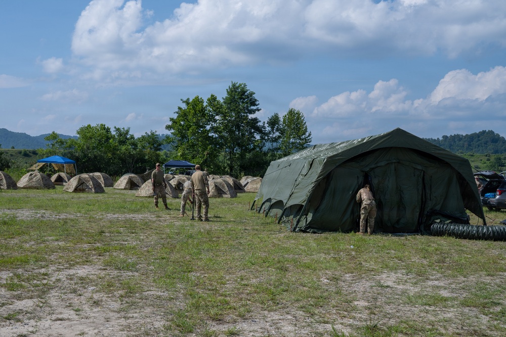 A military field camp is being established in an open grassy area surrounded by forested hills. Personnel in camouflage uniforms are setting up tents, including a large olive-drab command or medical tent in the foreground and multiple smaller camouflage individual tents in the background. A flexible ventilation duct is visible connected to the large tent, suggesting climate control or air circulation equipment.