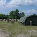 A military field camp is being established in an open grassy area surrounded by forested hills. Personnel in camouflage uniforms are setting up tents, including a large olive-drab command or medical tent in the foreground and multiple smaller camouflage individual tents in the background. A flexible ventilation duct is visible connected to the large tent, suggesting climate control or air circulation equipment.
