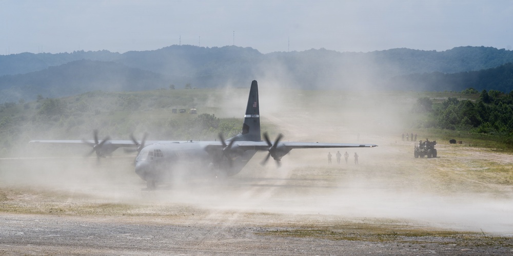 A military C-130 Hercules turboprop transport aircraft is operating on an unpaved or semi-prepared airstrip, generating large dust clouds from its propeller wash. Ground personnel and military vehicles are visible nearby, suggesting a tactical or expeditionary airfield operation. The scene appears to be a military exercise or real-world logistics mission in a hilly, vegetated environment.
