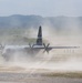 A military C-130 Hercules turboprop transport aircraft is operating on an unpaved or semi-prepared airstrip, generating large dust clouds from its propeller wash. Ground personnel and military vehicles are visible nearby, suggesting a tactical or expeditionary airfield operation. The scene appears to be a military exercise or real-world logistics mission in a hilly, vegetated environment.