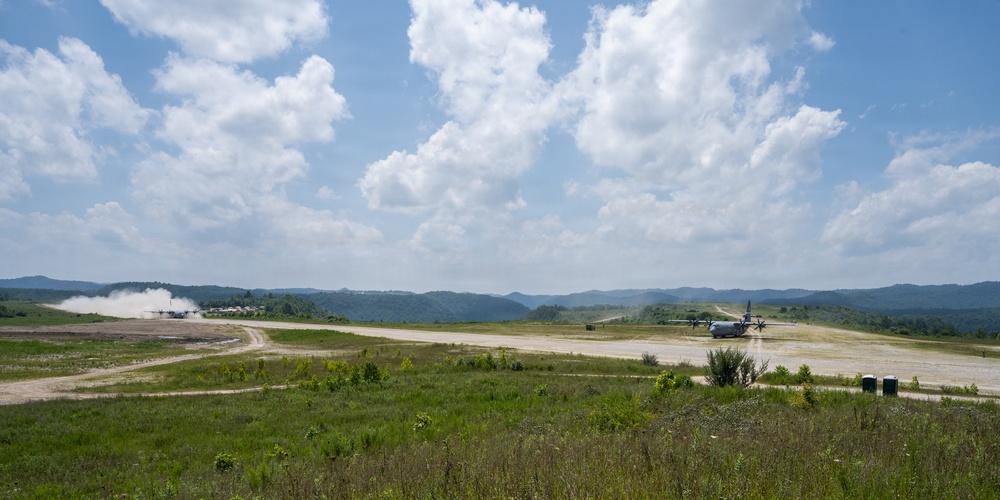 The image shows a military airstrip in a rural, hilly environment with two large turboprop aircraft visible on the runway. One aircraft on the left appears to be taking off or landing, generating a visible dust cloud, while a second aircraft is stationary or taxiing on the right side of the runway. The surrounding terrain features green rolling hills, sparse vegetation, and an overcast-to-partly-cloudy sky.