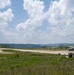 The image shows a military airstrip in a rural, hilly environment with two large turboprop aircraft visible on the runway. One aircraft on the left appears to be taking off or landing, generating a visible dust cloud, while a second aircraft is stationary or taxiing on the right side of the runway. The surrounding terrain features green rolling hills, sparse vegetation, and an overcast-to-partly-cloudy sky.