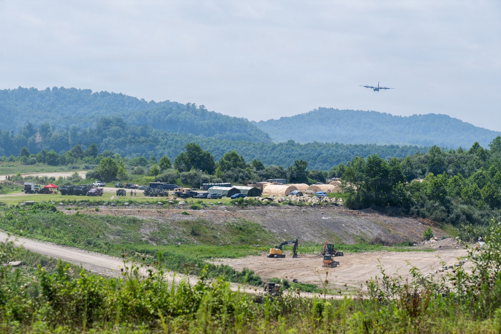 The image depicts a military forward operating base or temporary staging area situated in a valley surrounded by forested hills. A large military transport aircraft, consistent with a C-130 Hercules, is visible in flight approaching or departing the area. Construction equipment and military vehicles are actively present, suggesting ongoing site preparation or disaster relief operations.