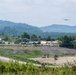 The image depicts a military forward operating base or temporary staging area situated in a valley surrounded by forested hills. A large military transport aircraft, consistent with a C-130 Hercules, is visible in flight approaching or departing the area. Construction equipment and military vehicles are actively present, suggesting ongoing site preparation or disaster relief operations.