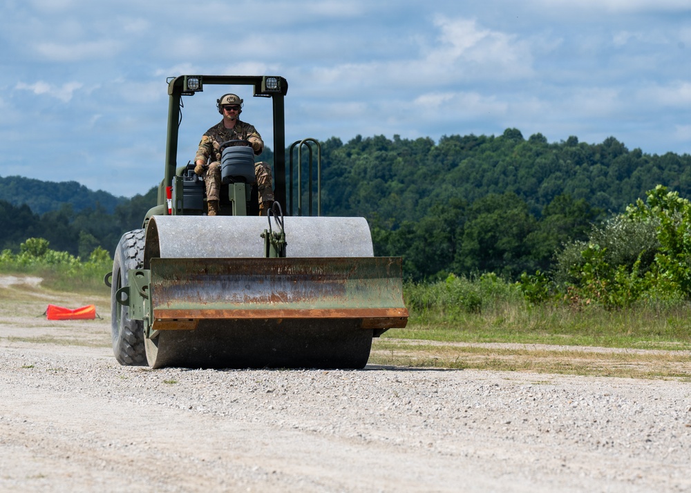 A military service member in camouflage uniform and tactical headgear operates a military-green road compactor/roller on a gravel surface. The heavy construction equipment features a large steel drum roller at the front and is being used to compact or grade a dirt/gravel road or airstrip. The setting appears to be a rural military training or construction site surrounded by dense tree lines.