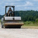 A military service member in camouflage uniform and tactical headgear operates a military-green road compactor/roller on a gravel surface. The heavy construction equipment features a large steel drum roller at the front and is being used to compact or grade a dirt/gravel road or airstrip. The setting appears to be a rural military training or construction site surrounded by dense tree lines.