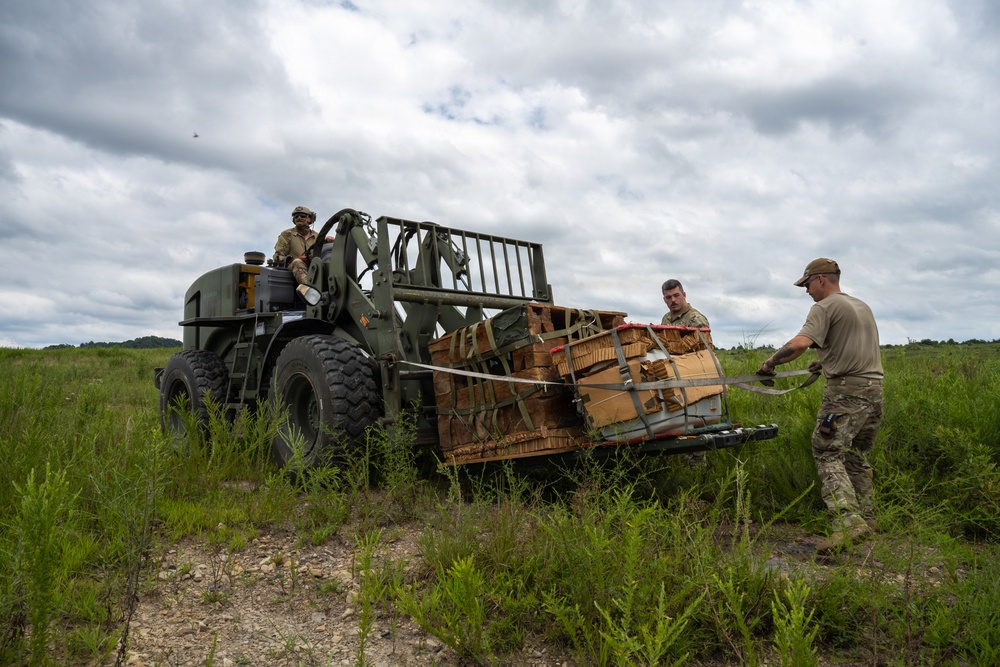 Military personnel are working with a military rough terrain forklift in an open grassy field. Three soldiers are handling what appears to be a cargo load secured with straps and netting on the forks of the vehicle. The scene appears to be a field logistics or resupply operation, likely during a training exercise.