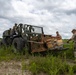 Military personnel are working with a military rough terrain forklift in an open grassy field. Three soldiers are handling what appears to be a cargo load secured with straps and netting on the forks of the vehicle. The scene appears to be a field logistics or resupply operation, likely during a training exercise.