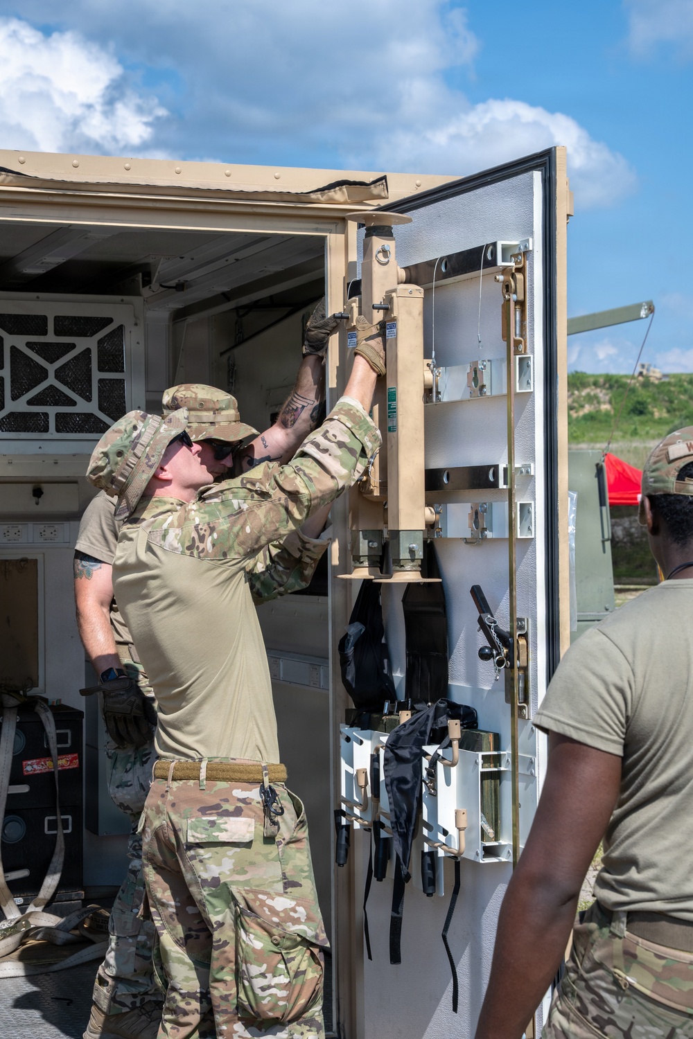 Military personnel in OCP (Operational Camouflage Pattern) uniforms are working on the door mechanism of a tan/desert-colored military shelter or vehicle enclosure. The primary soldier is reaching upward to manipulate locking hardware on a large equipment door. The setting appears to be an outdoor military training or operational area under partly cloudy skies.