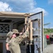Military personnel in OCP (Operational Camouflage Pattern) uniforms are working on the door mechanism of a tan/desert-colored military shelter or vehicle enclosure. The primary soldier is reaching upward to manipulate locking hardware on a large equipment door. The setting appears to be an outdoor military training or operational area under partly cloudy skies.