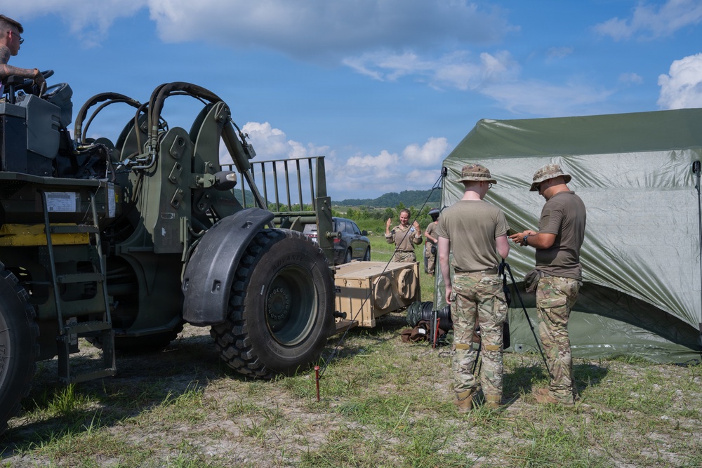 Military personnel are engaged in field operations, working near a large military forklift/rough terrain forklift and a green military tent structure. Soldiers in MultiCam/OCP uniforms and boonie hats appear to be setting up or managing equipment and rigging lines connected to the tent. The scene takes place on an open grassy field under partly cloudy skies, suggesting a field training exercise or forward operating base setup.