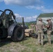 Military personnel are engaged in field operations, working near a large military forklift/rough terrain forklift and a green military tent structure. Soldiers in MultiCam/OCP uniforms and boonie hats appear to be setting up or managing equipment and rigging lines connected to the tent. The scene takes place on an open grassy field under partly cloudy skies, suggesting a field training exercise or forward operating base setup.