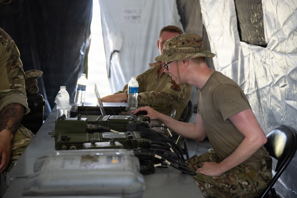 Military personnel are working inside a field tent at what appears to be a tactical operations setup. A soldier in the foreground wearing a boonie hat and glasses is operating military communications or electronic equipment on a folding table. A second soldier in the background is working on a laptop computer.
