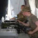 Military personnel are working inside a field tent at what appears to be a tactical operations setup. A soldier in the foreground wearing a boonie hat and glasses is operating military communications or electronic equipment on a folding table. A second soldier in the background is working on a laptop computer.