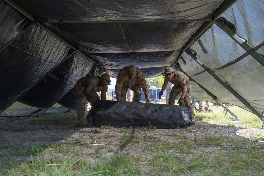 Three military personnel in camouflage uniforms are working together to set up or dismantle a large military tent structure. They are handling a rolled or folded black canvas/tarp component underneath the partially erected tent frame. The setting appears to be an outdoor field environment with gravel and grass ground cover.
