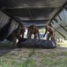 Three military personnel in camouflage uniforms are working together to set up or dismantle a large military tent structure. They are handling a rolled or folded black canvas/tarp component underneath the partially erected tent frame. The setting appears to be an outdoor field environment with gravel and grass ground cover.