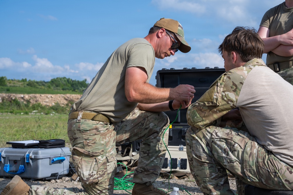 Two military personnel in camouflage uniforms are crouched down working with cables and equipment in an outdoor field setting. A third individual stands partially visible in the background. They appear to be setting up or troubleshooting electronic or communications equipment using a ruggedized case system.