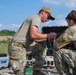 Two military personnel in camouflage uniforms are crouched down working with cables and equipment in an outdoor field setting. A third individual stands partially visible in the background. They appear to be setting up or troubleshooting electronic or communications equipment using a ruggedized case system.